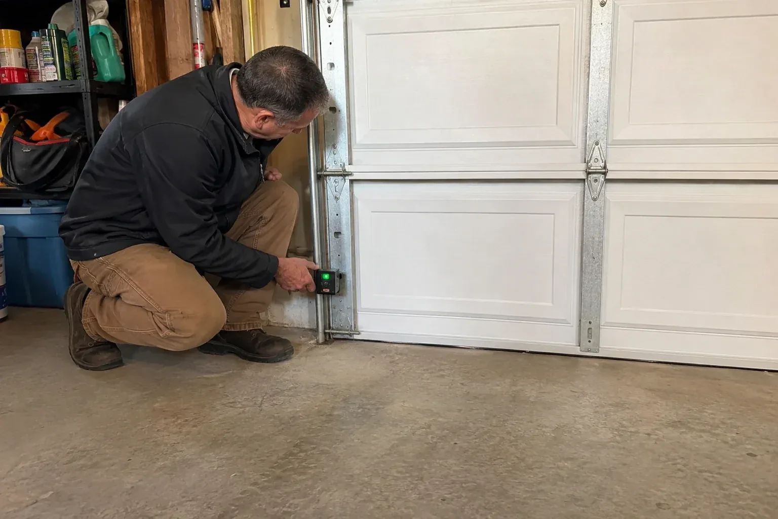 Technician kneeling beside a white paneled garage door, holding a small black box with a green light against the vertical track. — Photo-Eye Sensor Alignment or Replacement, Vaughan