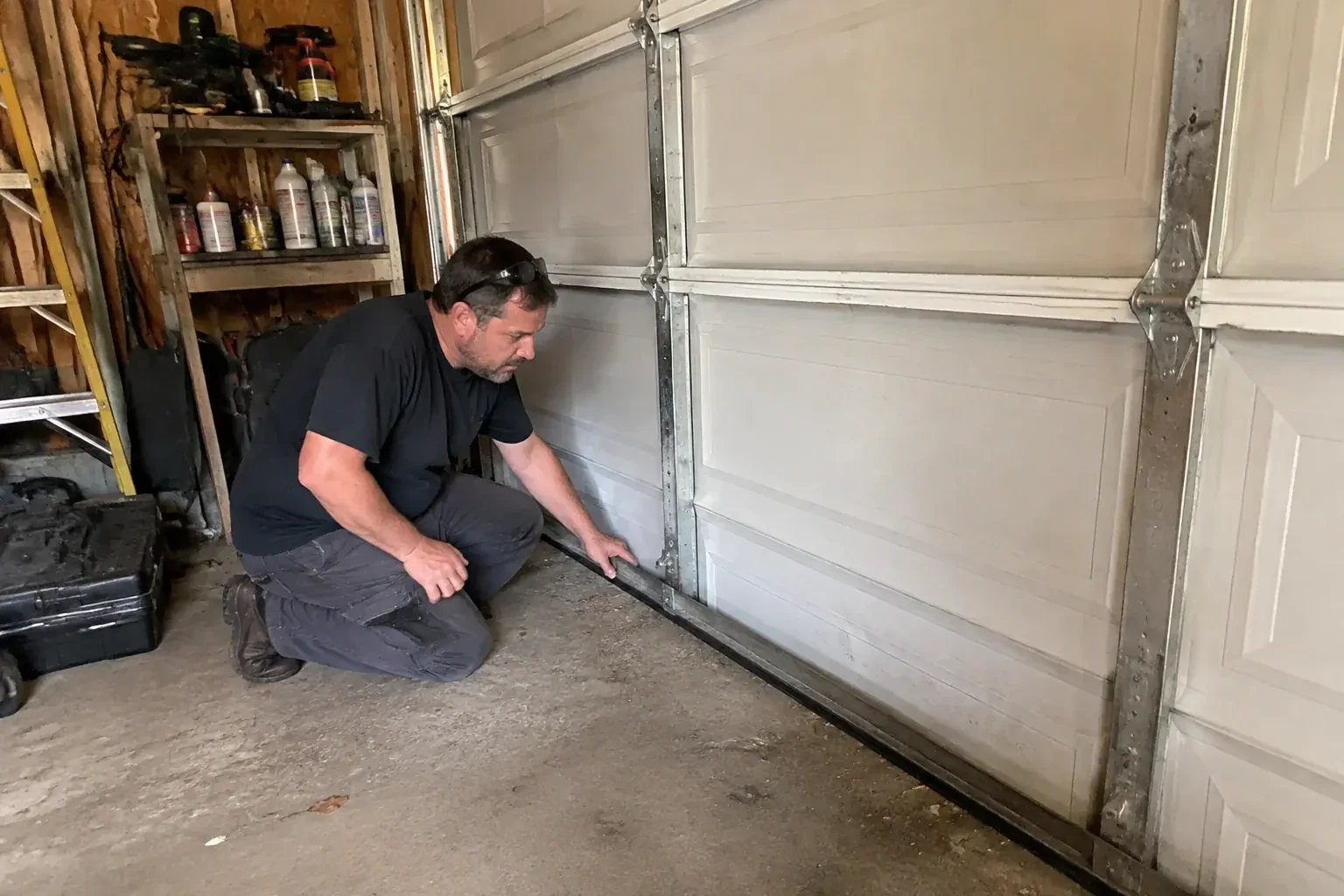 Person kneeling by a white paneled door in a garage, one hand on the black seal along its bottom edge. — Garage Door Roller Replacement, Vaughan